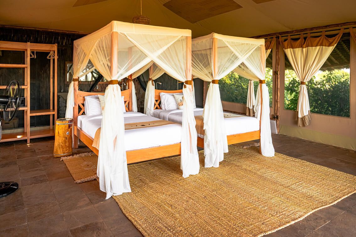 A four-poster bed with white linens and a woven rug, set in a room with large windows and a view of greenery.