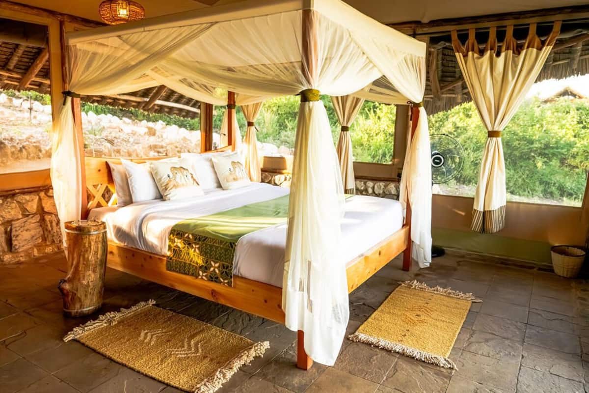 Uma cama de dossel com cortinas claras, roupa de cama branca, cabeceira de madeira e vista para o verde lá fora.