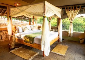 Una cama con dosel y cuatro columnas, con sábanas blancas, cabecera de madera y vistas al exterior con vegetación.