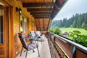 A wooden balcony with a table and chairs, a railing, and a view of a forested landscape.