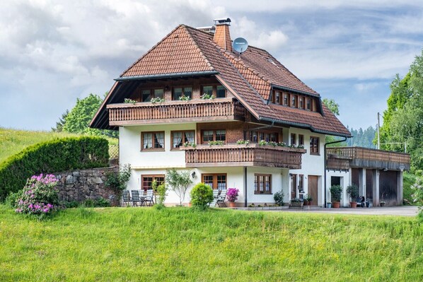 A two-story house with a sloped roof, wooden balconies, and a stone wall.