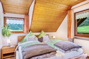 A bedroom with a sloped ceiling, a bed with multiple pillows, a bedside table with a lamp, and a window with curtains.