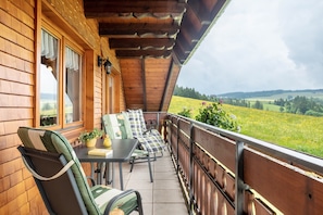 A wooden balcony with a table and chairs, a view of a green field, and a cloudy sky.