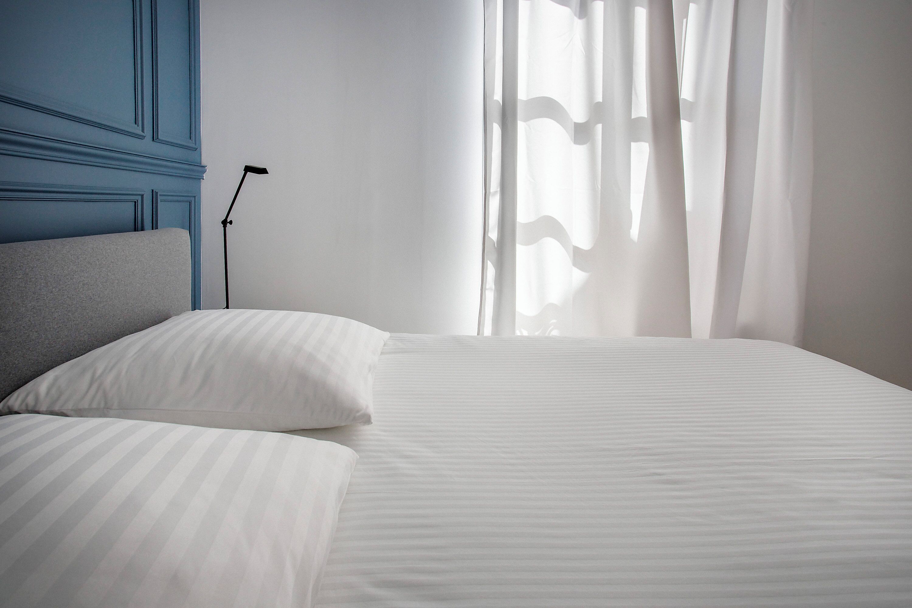 A neatly made bed with white bedding, a grey headboard, and a bedside lamp.