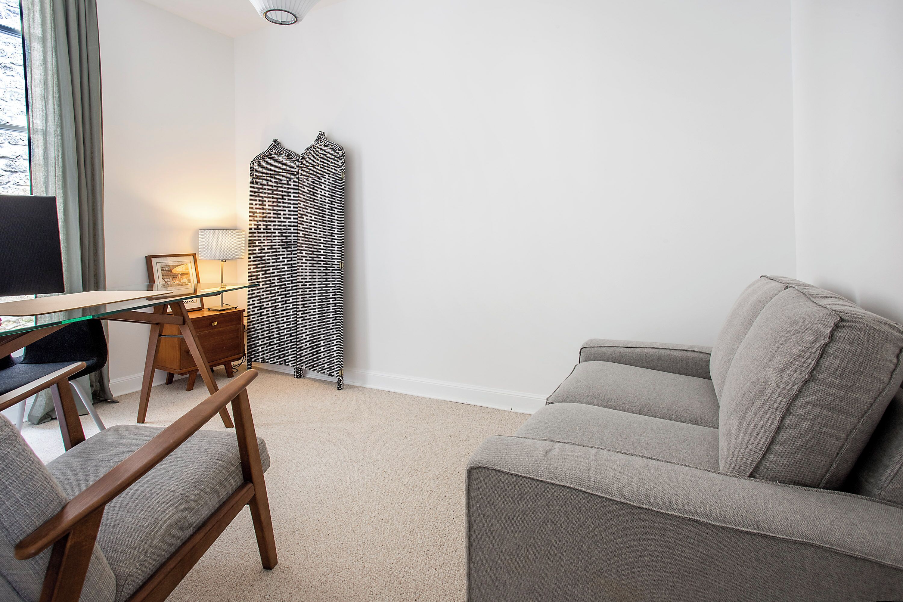 A modern living room with a glass-top desk, a grey sofa, a wooden chair, a lamp, and a patterned curtain.