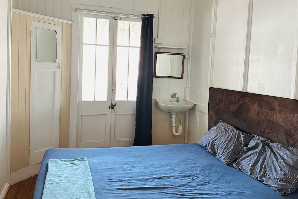 A bedroom with a bed, a ceiling fan, a window with curtains, a door, a small sink, and a mirror.