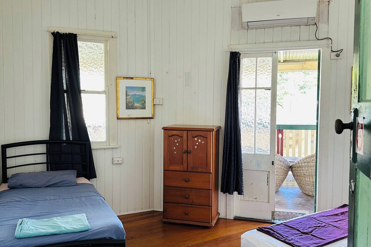 A bedroom with two beds, wooden wardrobe, white walls, ceiling fan, and a door leading outside.
