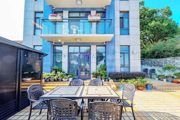 Un espacio exterior con mesas y sillas, un edificio con balcón y plantas en macetas.