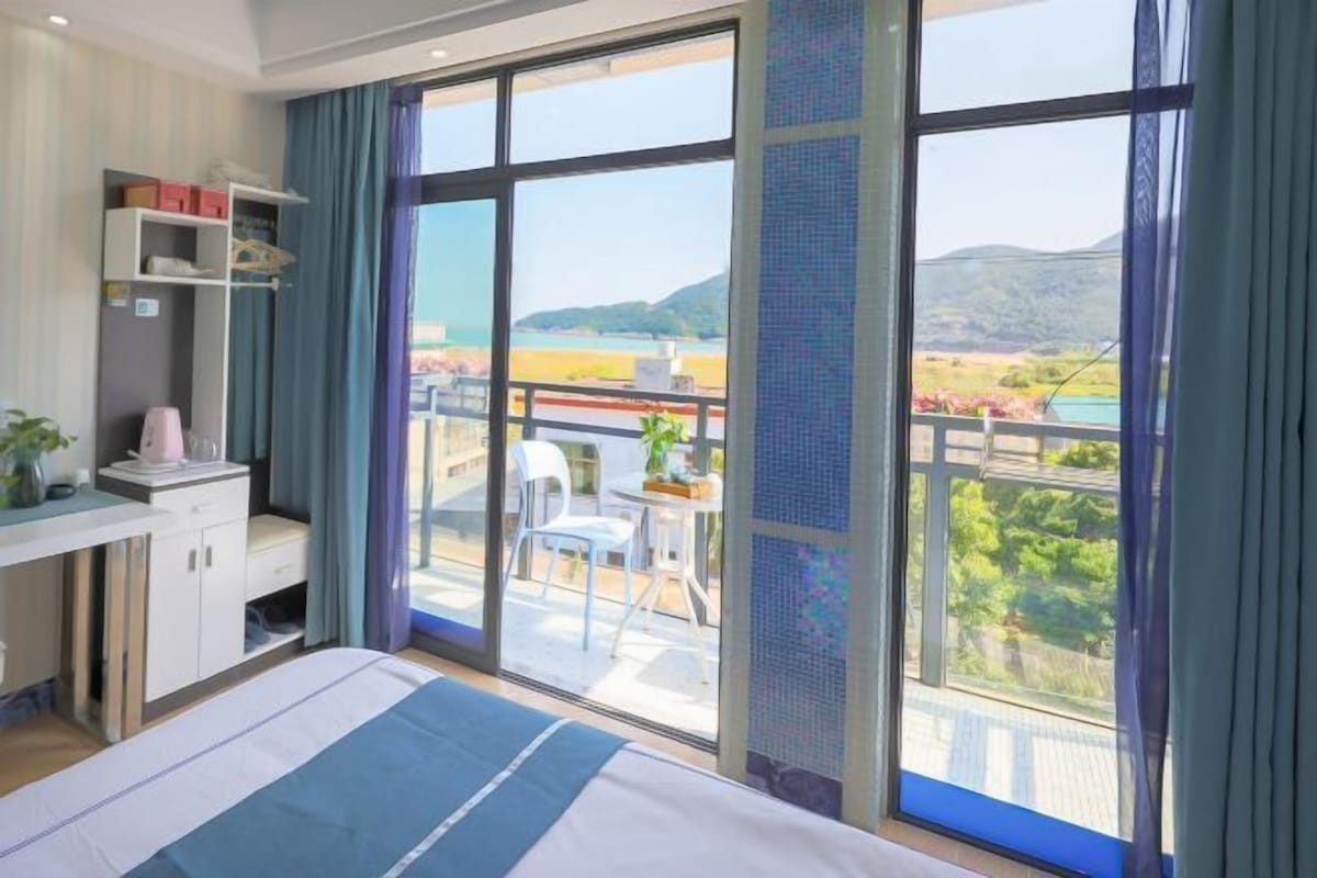 A hotel room with a large bed, a desk with a chair, a wardrobe, and a balcony with a view of the sea and hills.