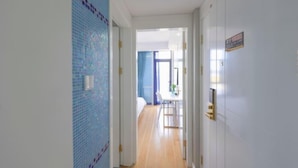 Un pasillo con pared de azulejos, una puerta con un cartel y vista a una habitación con cama y mesa.