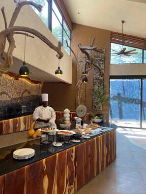 A chef at a wooden counter with a rustic design, surrounded by various food items and a decorative wall with geometric patterns. - Soroi Samburu Lodge (Samburu)