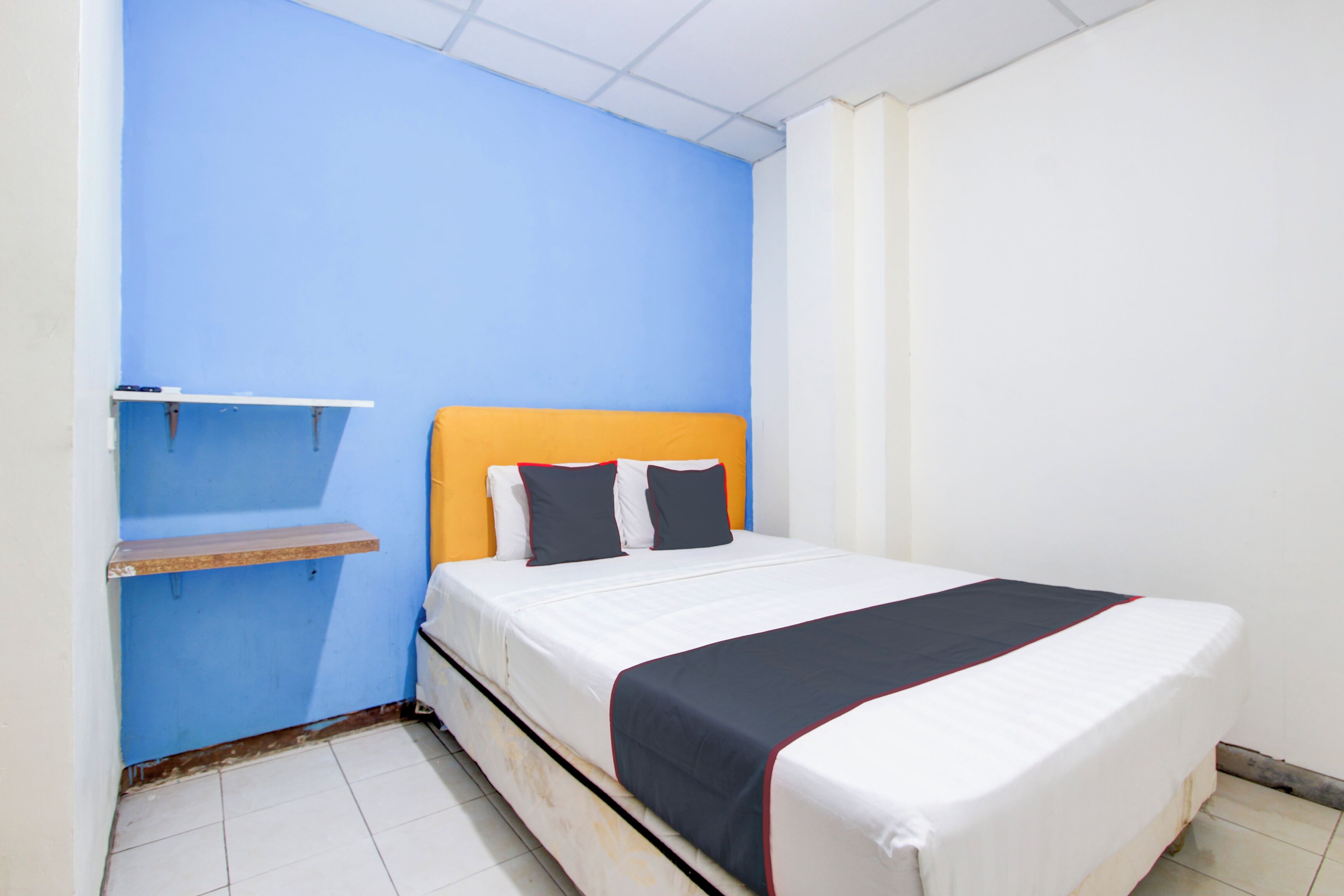 A small, single-bed room with a blue wall, a wooden shelf, and a white mattress with a navy and red accent.