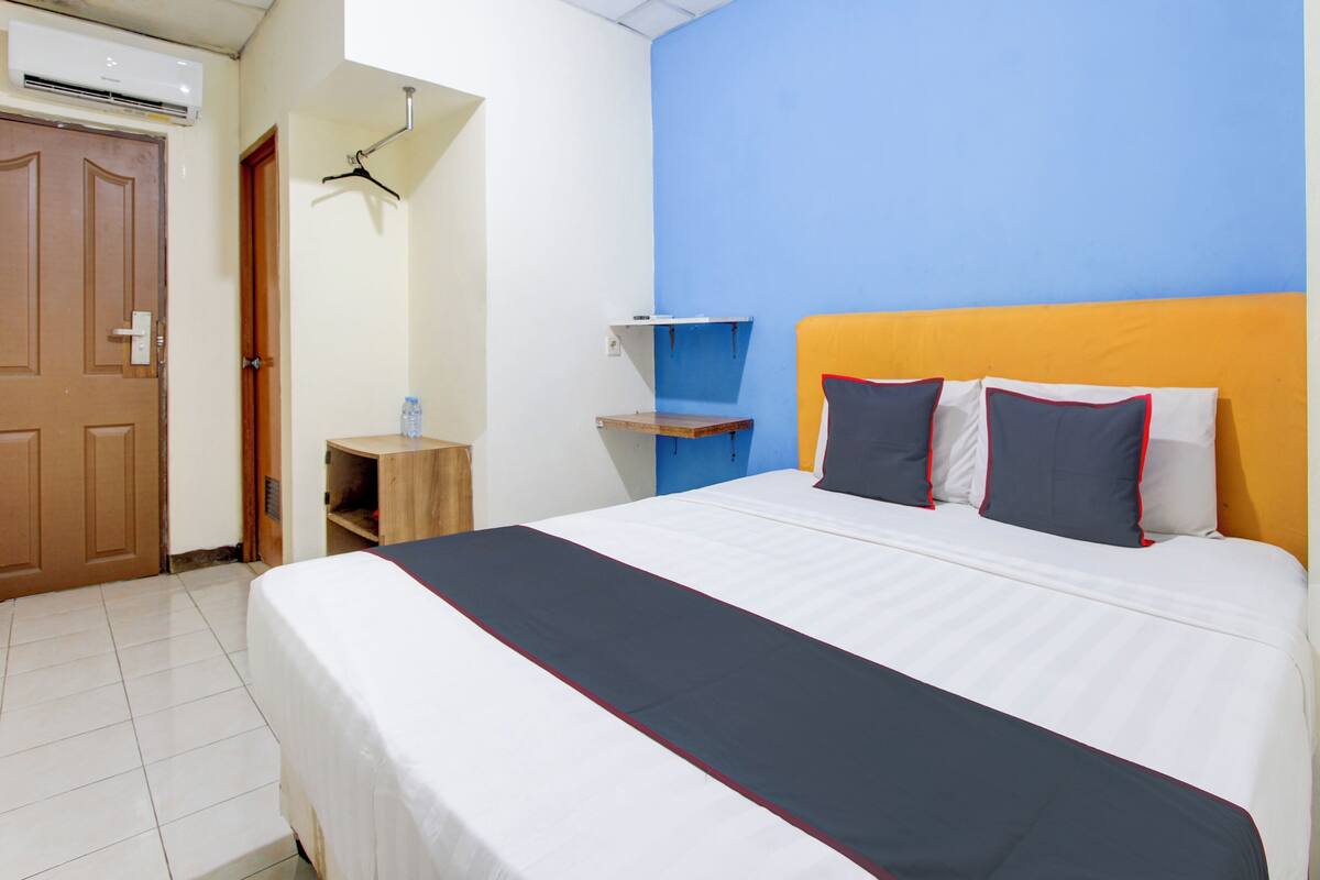 A hotel room with a bed, a wooden nightstand, a water bottle, a hanging coat rack, and a door.