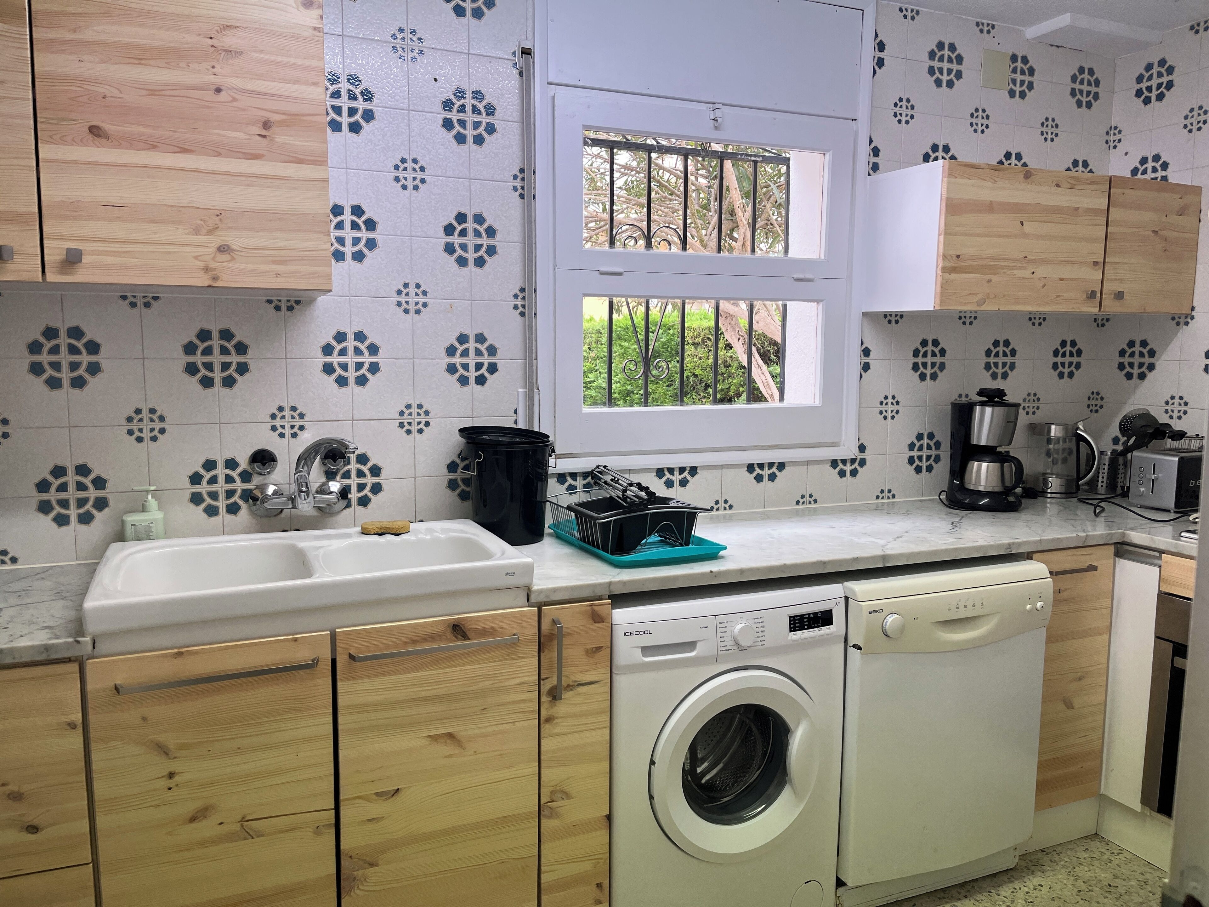 A kitchen with wooden cabinets, a washing machine, a coffee maker, and a window with a view of greenery.