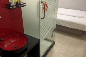 A bathroom with a red sink, a shower with a glass door, and a bed with white bedding.