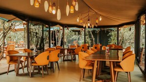 Een eetruimte binnenshuis met houten tafels en oranje stoelen, onder een grote tent met hangende lampen.