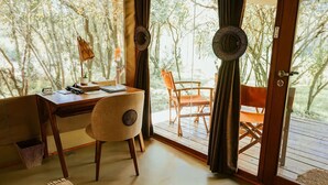 Een kamer met een houten bureau, een stoel, een klein tafeltje met een plant, en uitzicht op een terras en bomen.