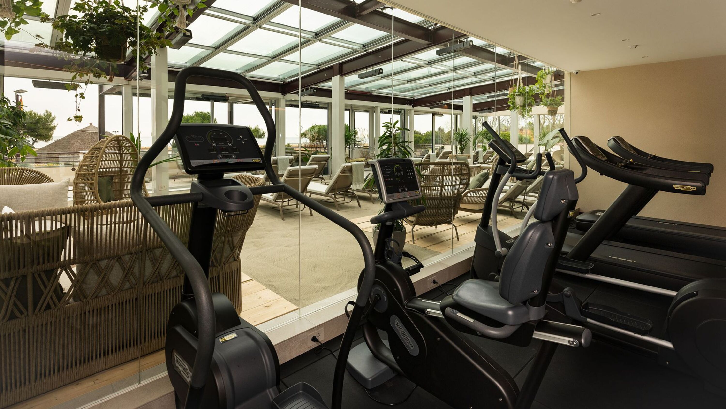 Un gimnasio con bicicletas estáticas y cinta de correr, ventanas amplias y zona de estar al aire libre.