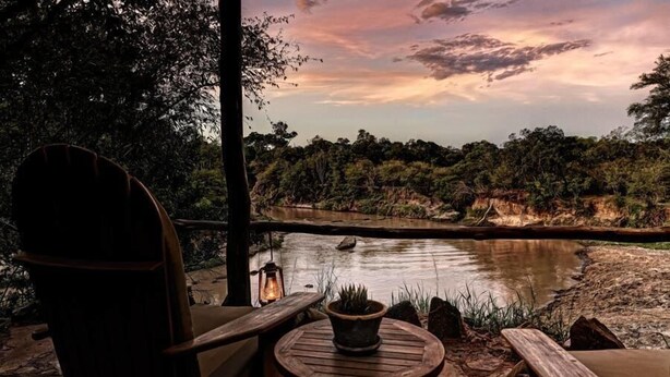 A wooden deck with a chair and a table, overlooking a river with a forested landscape and a sunset sky. - Soroi Private Camp (Maasai Mara)