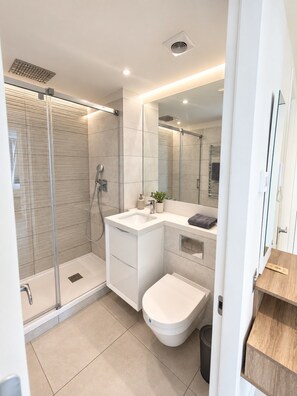 Un bany modern amb plat de dutxa envoltat per vidre, un lavabo blanc i un vàter.