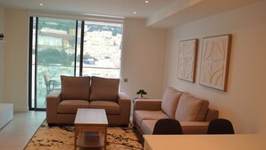 Een moderne woonkamer met een bank, een salontafel en een eettafel. Er is een groot raam met uitzicht op gebouwen.