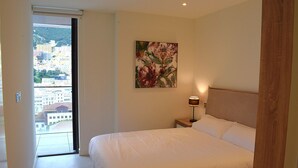 Een slaapkamer met een bed, nachtkastje, lamp en schilderij. Er is een raam met uitzicht op gebouwen en een deur.
