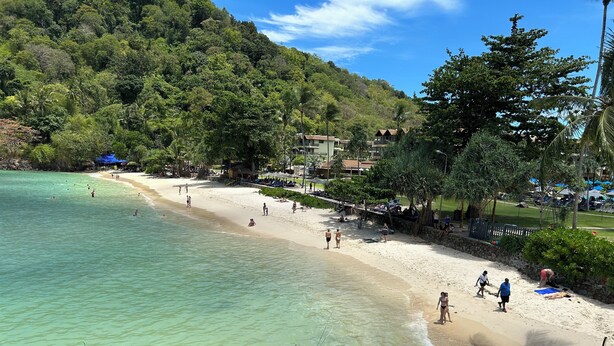 Ein Strand mit klarem türkisblauem Wasser, einem sandigen Ufer und üppiger Vegetation im Hintergrund.