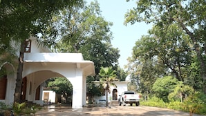A paved pathway leading to a white building with an arched entrance, surrounded by lush greenery and a parked vehicle.