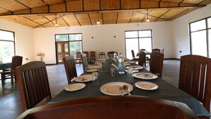 A dining area with a long table set for a meal, surrounded by wooden chairs, with a thatched ceiling and large windows.