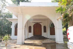 A white building with a central wooden door and arched windows, surrounded by a paved area and palm trees. - Bamera House Jungle Retreat Bandhavgarh (Umaria)
