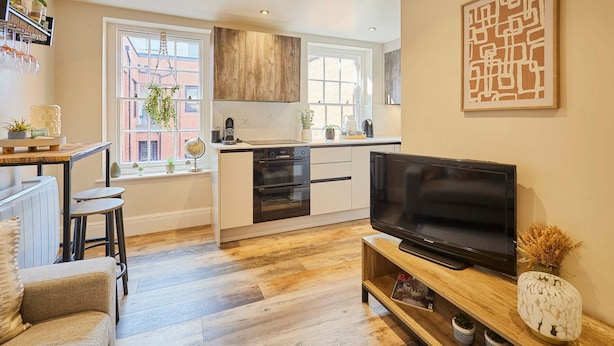 Una cocina moderna con horno empotrado, una zona de comedor con mesa y sillas, y un televisor sobre un mueble de madera.