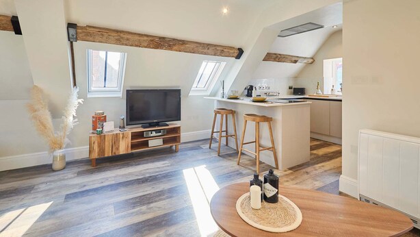 Eine moderne Loftswohnung mit Küche, Essbereich und Wohnbereich. Sie zeichnet sich durch freiliegende Holzbalken, ein großes Fenster und einen Flachbildfernseher aus.