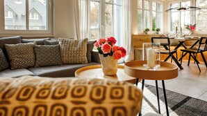A well-lit living room with a sofa, a coffee table with a vase of flowers, and a dining area with chairs and a table. - Lieblingshof House 4 - Holidays between the Baltic Sea and the Bodden (Born a. Darß)