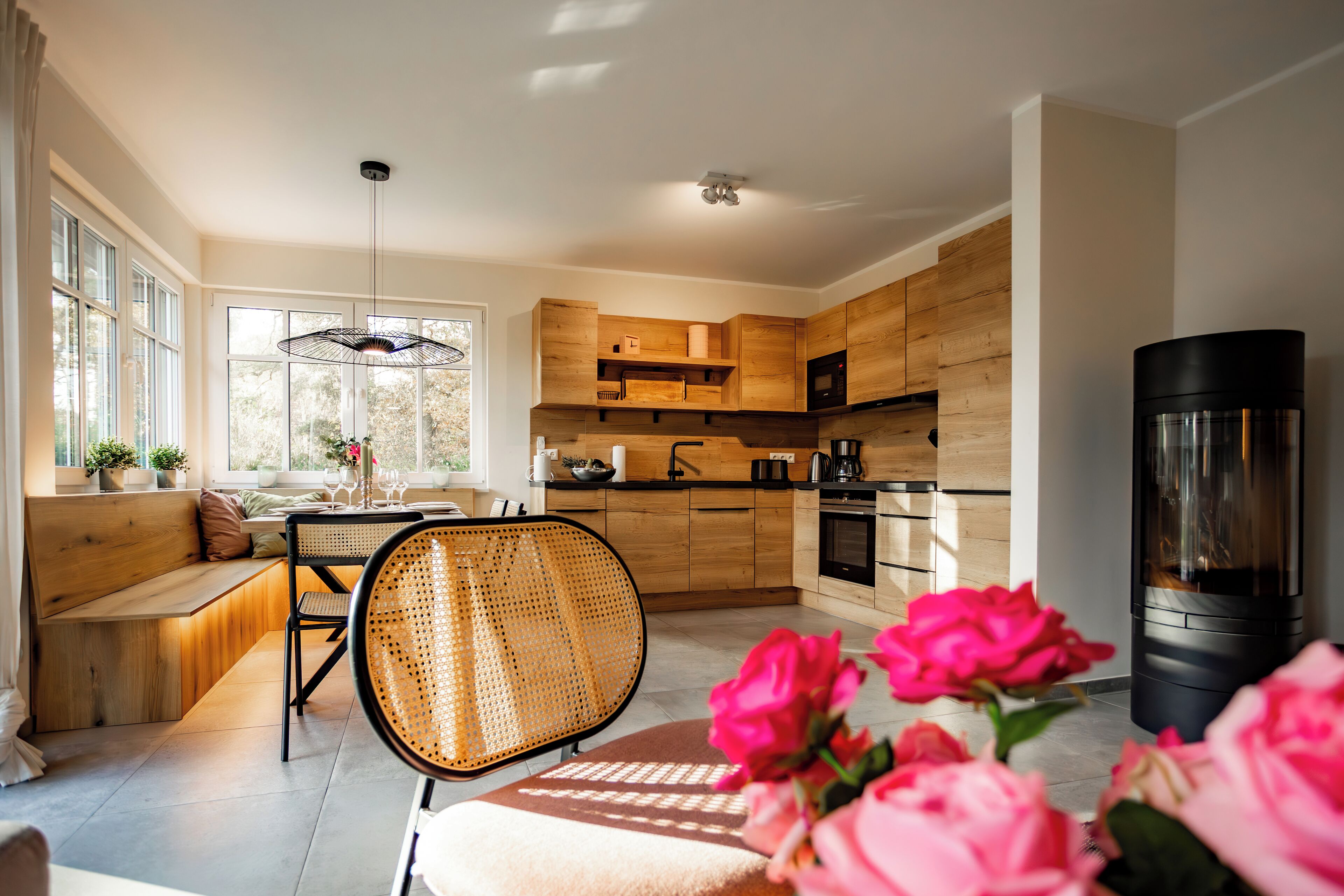 A modern kitchen with wooden cabinets, a black stove, and a dining area with a bench and rattan chair.