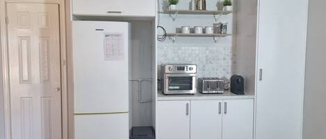 A compact kitchen with a refrigerator, oven, toaster, and shelves with decorative items.