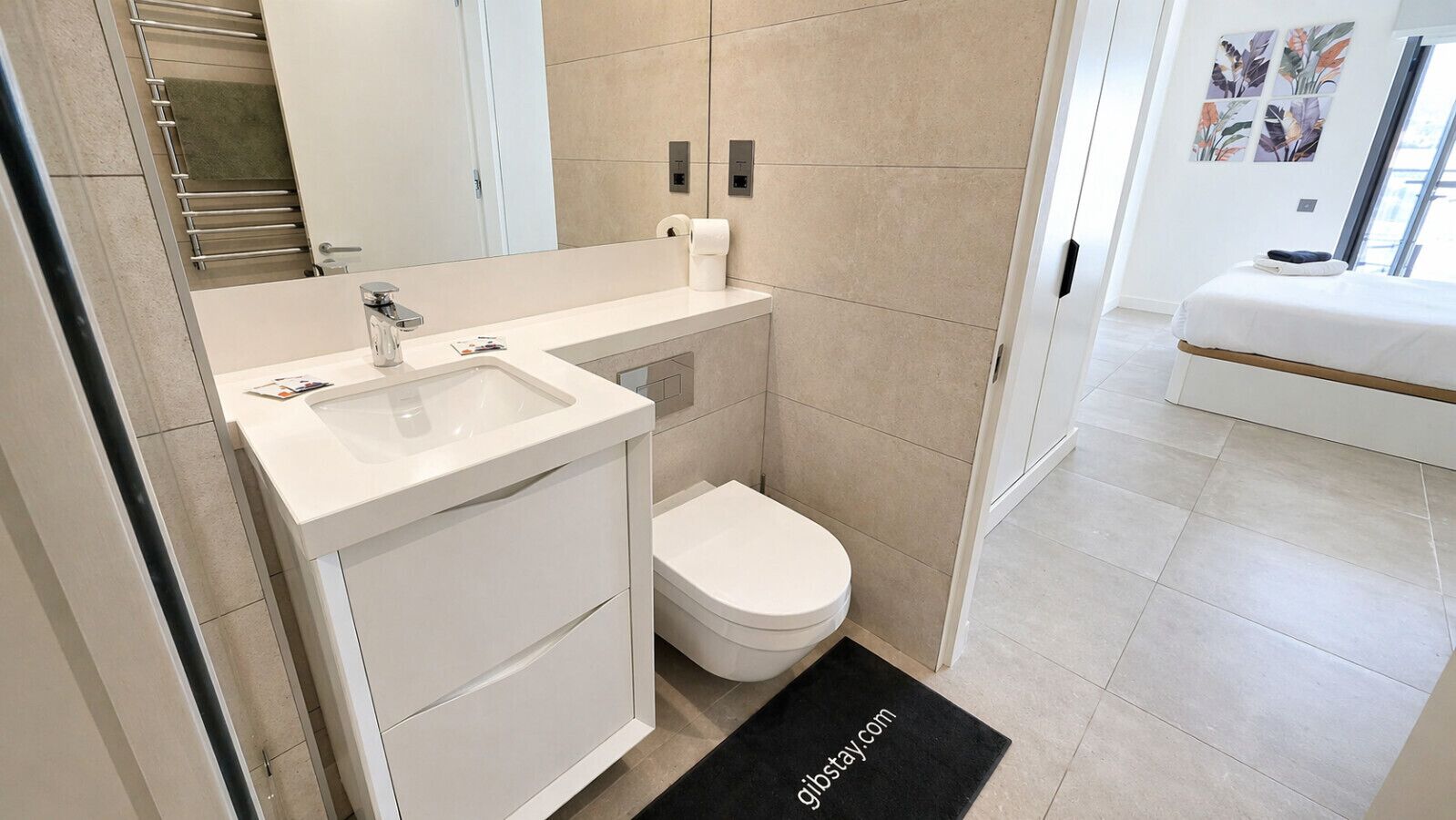 Un baño moderno con un lavamanos, inodoro y tocador blancos. Se observa un felpudo negro con texto, y un tendedero para toallas.