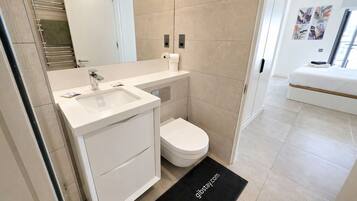 Un baño moderno con un lavamanos, inodoro y tocador blancos. Se observa un felpudo negro con texto, y un tendedero para toallas.