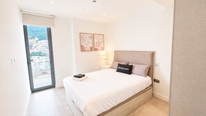 Ein Schlafzimmer mit Bett, Nachttisch, Lampe und Blick auf eine Stadtlandschaft durch eine Glastür.