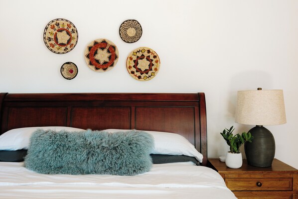 Een netjes opgemaakt bed met een zacht kussen, een houten nachtkastje met een lamp en een plant, en een wand met decoratieve wandborden.