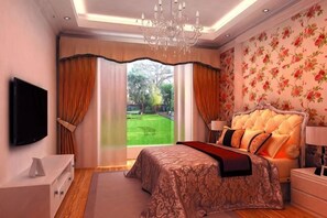 Een slaapkamer met een groot bed, een televisie, een kroonluchter en uitzicht op een tuin.