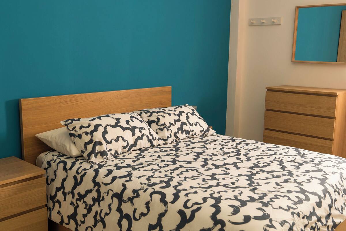 Un llit amb un estovat estampat, un capçal de fusta i una tauleta de nit amb calaixos.