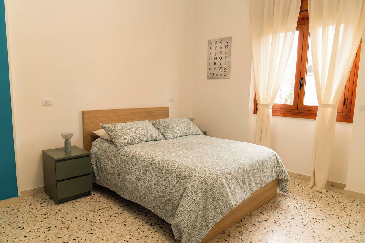 Un dormitori amb un llit, una taula de nit, un rellotge de paret i una finestra amb cortines.