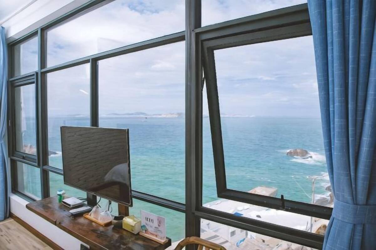Et værelse med stort vindue der vender ud mod havet, et skrivebord med en computer, og udsigt til kystlinjen.