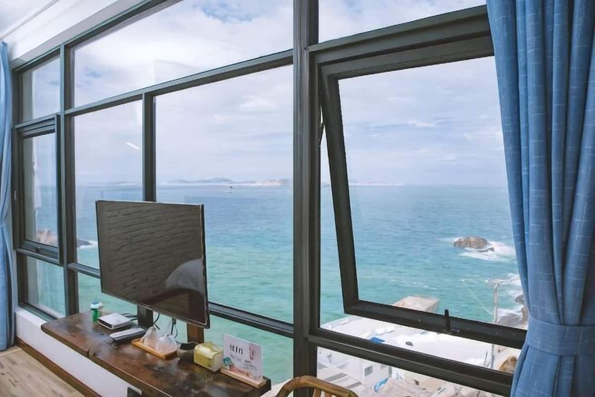 A room with a large window overlooking the sea, a desk with a computer, and a view of the coastline.