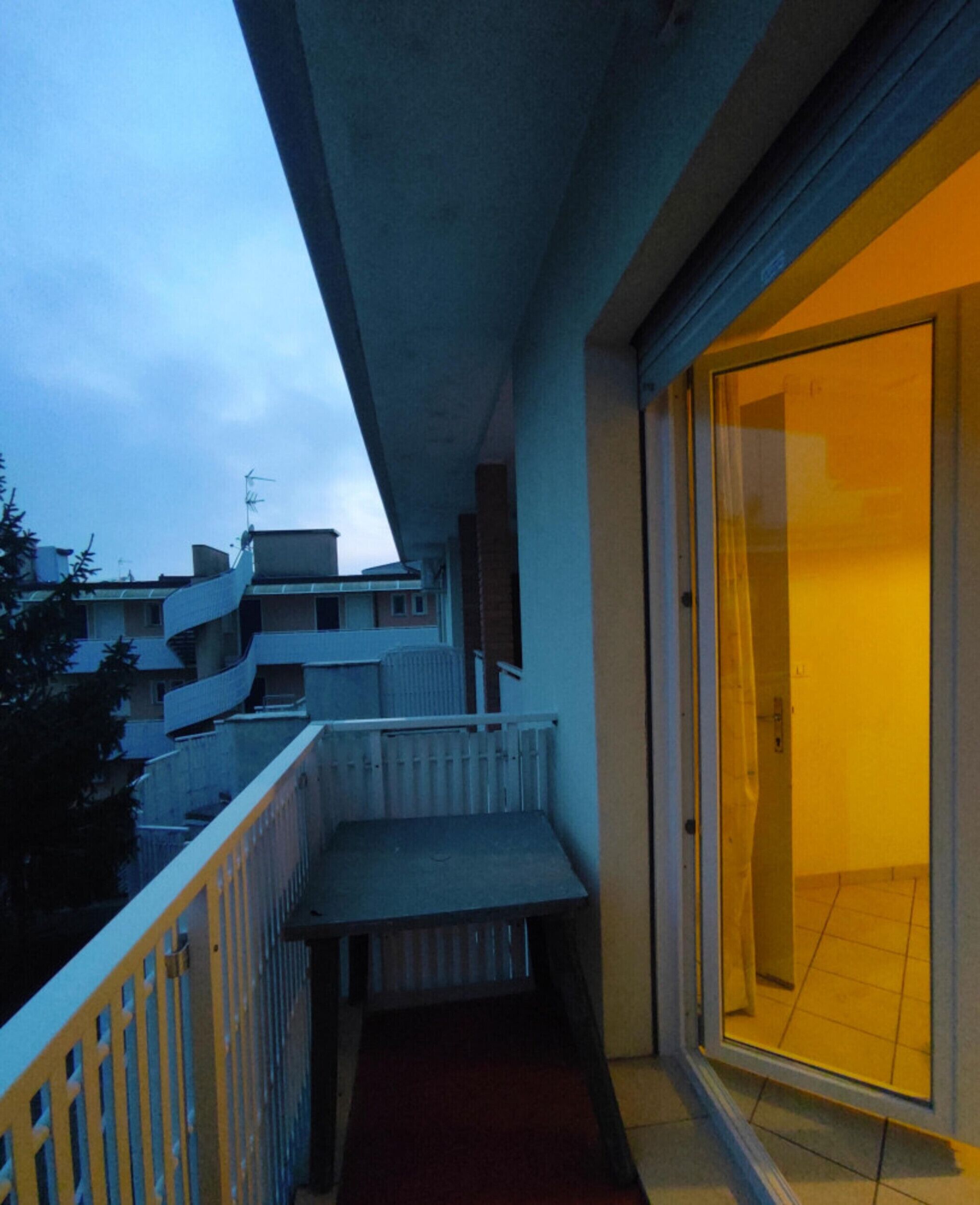 A narrow balcony with a yellow railing, a red mat, and a view of neighboring buildings.