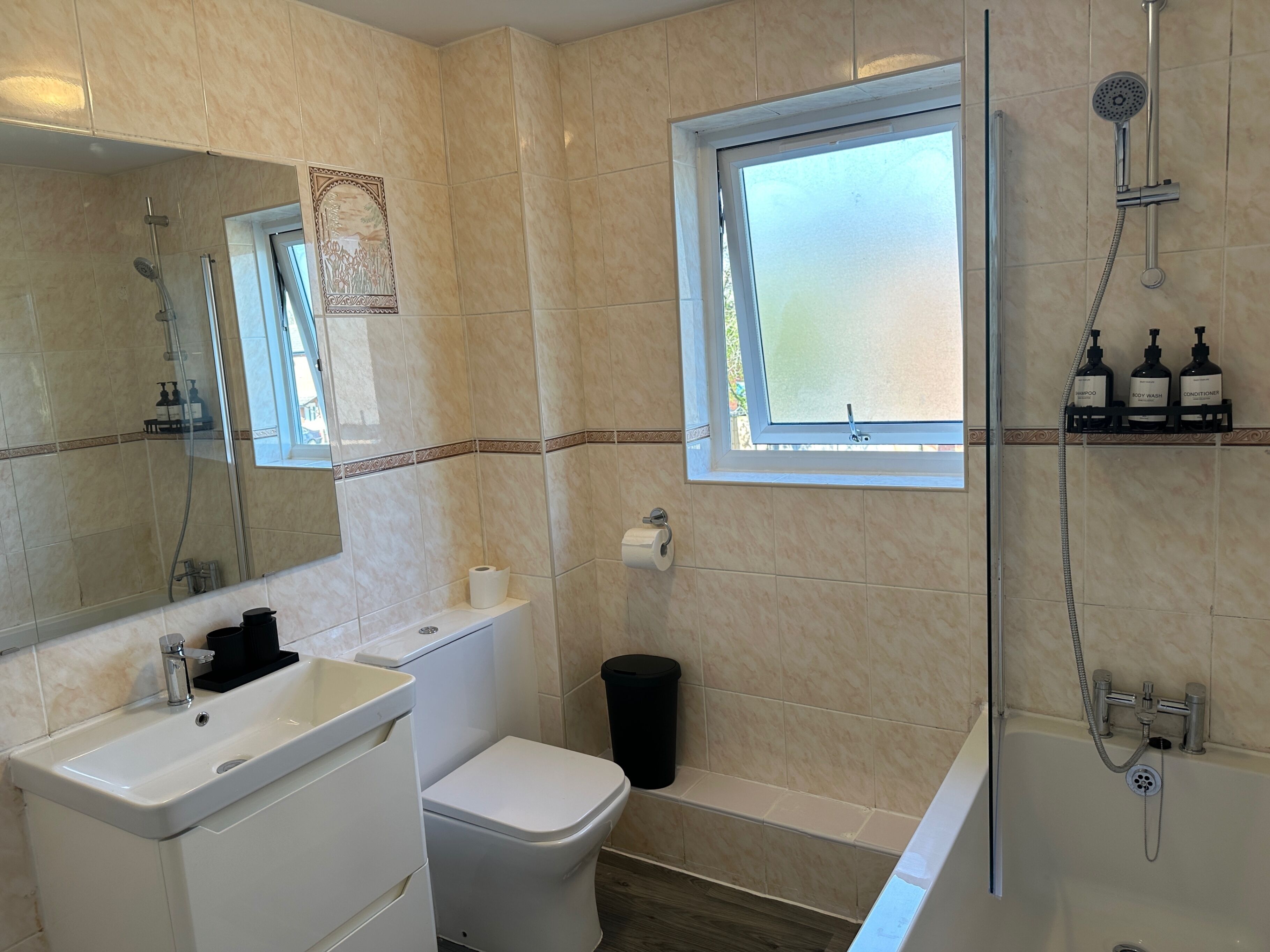 A bathroom with a toilet, sink, mirror, shower, and a window.