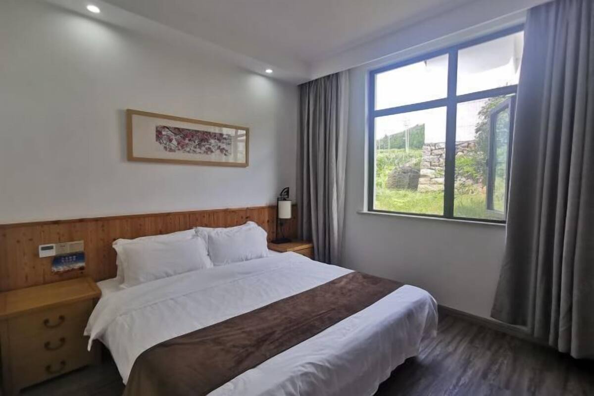 Habitación de hotel con una cama grande, cabecera de madera, mesita de noche, un cuadro enmarcado en la pared y una ventana con cortinas.