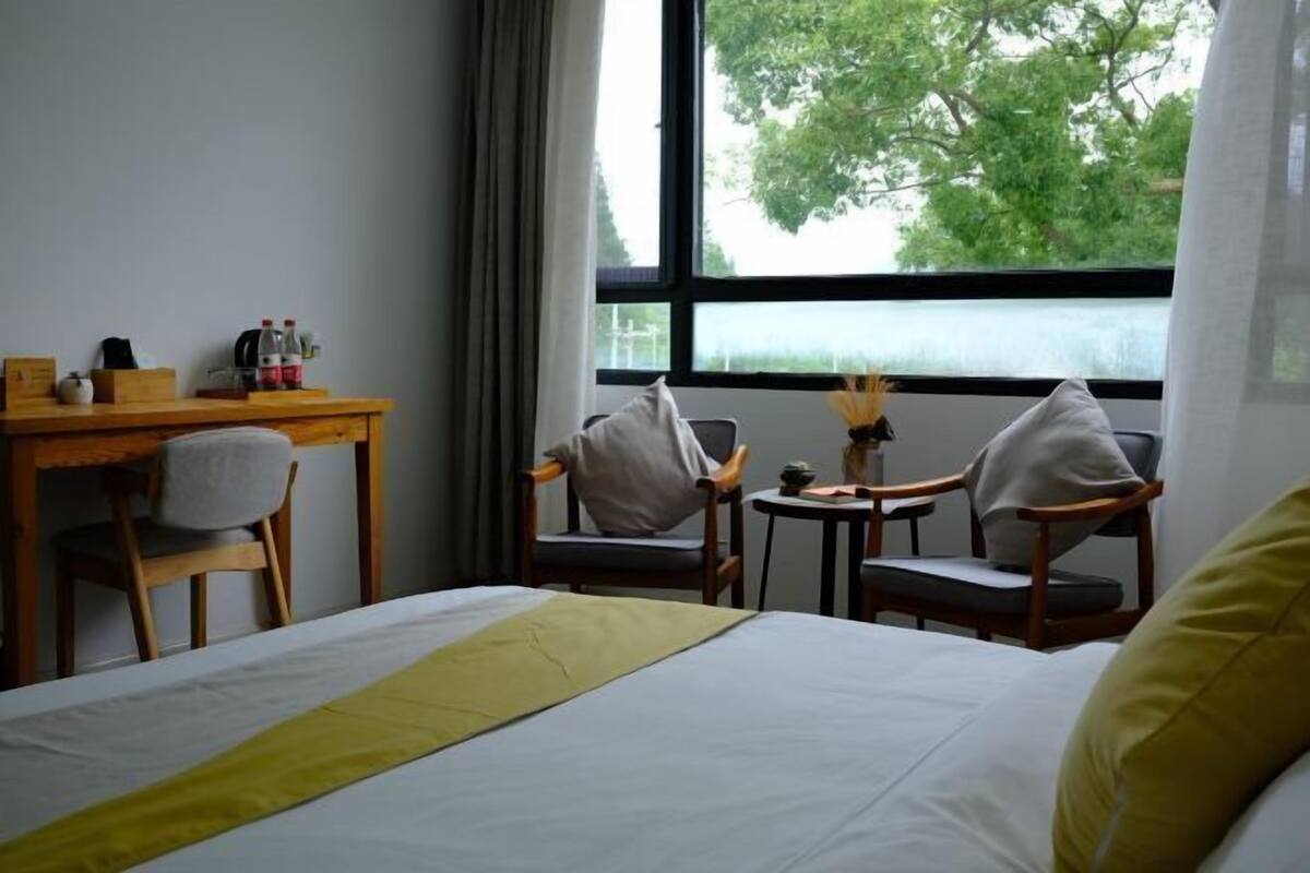Habitación de hotel con cama, dos sillas, una mesita y ventana con vista a árboles.