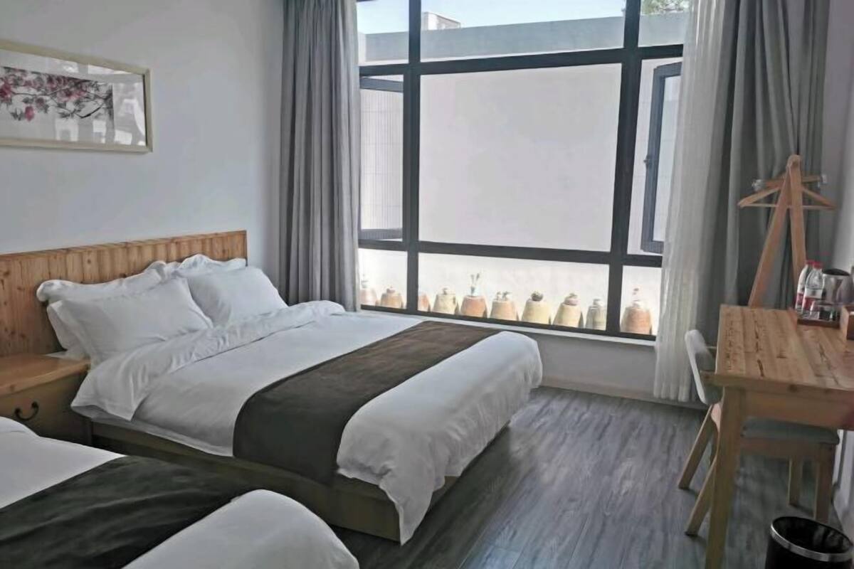 Una habitación de hotel con un ventanal grande, una cama de madera, un escritorio de madera y una mesita pequeña.