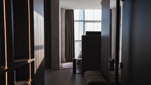 A modern hotel room with a dark wood floor, a large window with sheer curtains, a seating area with a sofa and a small table, and a wall-mounted shelf with LED lights. - DISTRICT boutique  (Osijek)